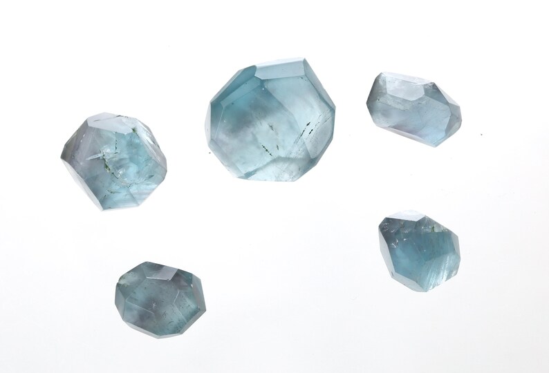 May include: Five faceted blue-green crystals, each with a unique shape and size, are displayed on a white background.