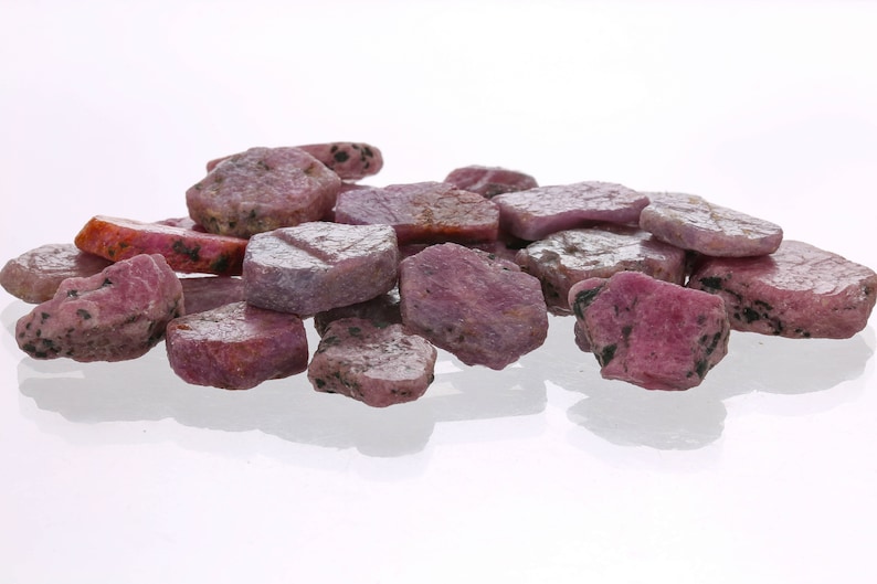 May include: A pile of pink ruby rough gemstones. The rough gemstones are various shapes and sizes. The gemstones are on a white background.