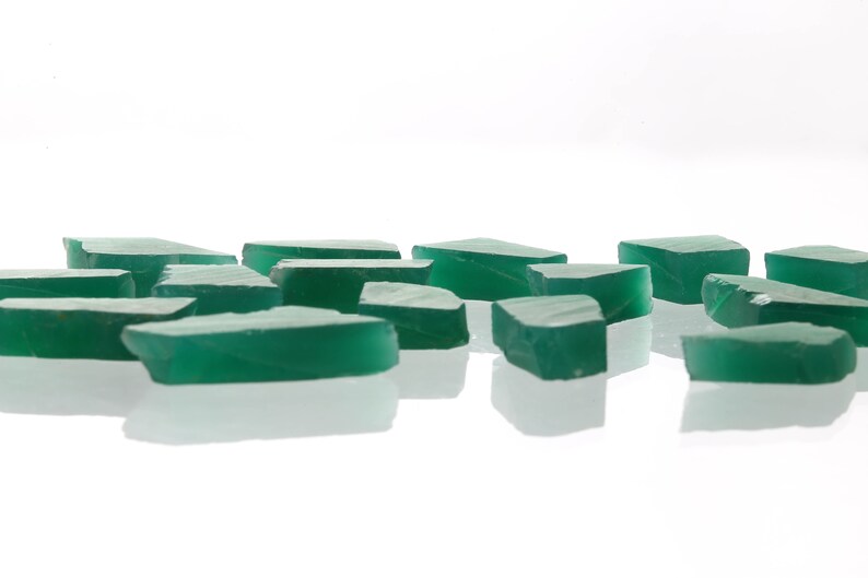 May include: A collection of green, irregularly shaped, translucent glass pieces. The pieces are scattered on a white surface.