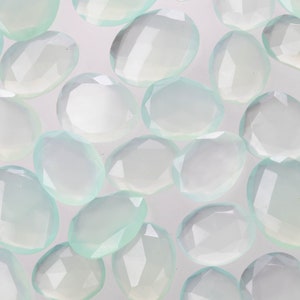 Puede incluir: Una colecci&oacute;n de piedras preciosas ovaladas facetadas de color verde p&aacute;lido. Las piedras est&aacute;n dispuestas en un patr&oacute;n aleatorio sobre un fondo blanco.