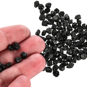 May include: A hand holding a small pile of black charcoal pieces. A larger pile of black charcoal pieces is scattered on a white surface.
