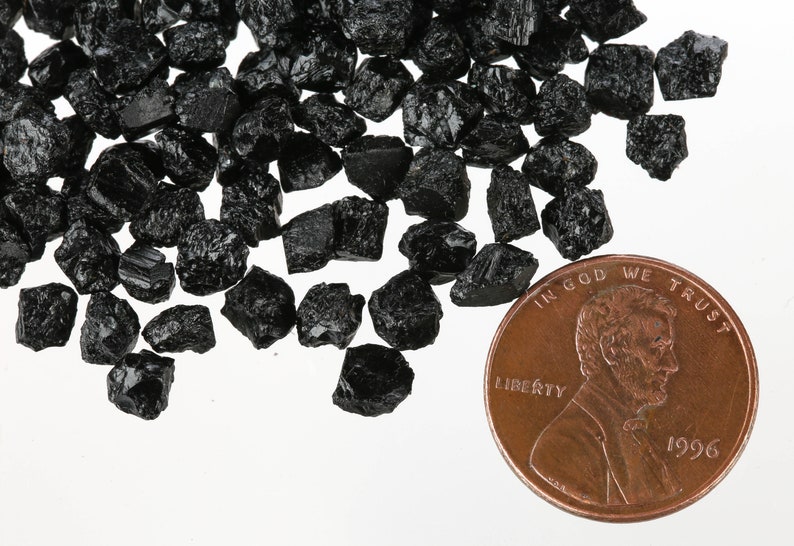 May include: A pile of black, rough, and jagged black tourmaline crystals next to a US penny for scale.  The crystals are about the size of a penny.