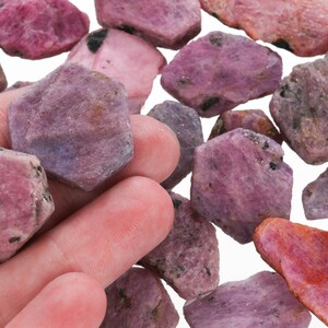 May include: A collection of rough pink and purple gemstones, likely rubies, with a variety of shapes and sizes. The stones have a natural, unpolished look.