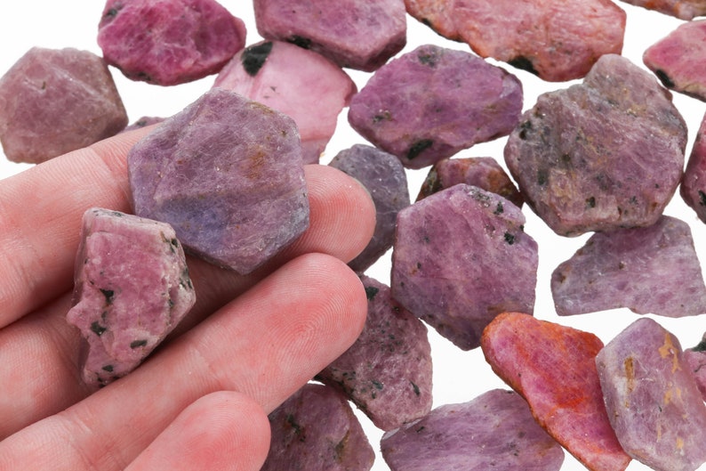 May include: A collection of rough pink and purple gemstones, likely rubies, with a variety of shapes and sizes. The stones have a natural, unpolished look.