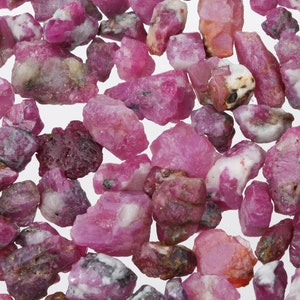 Peut inclure: Une collection de pierres précieuses de rubis roses, certaines avec des inclusions blanches, éparpillées sur une surface blanche.