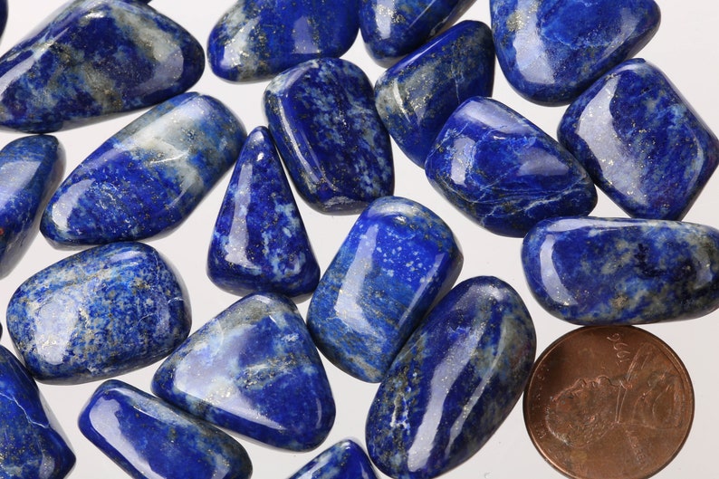 May include: A collection of tumbled lapis lazuli stones, each with a unique blue and white pattern. The stones are smooth and polished, with a glossy finish. A penny is included for scale.