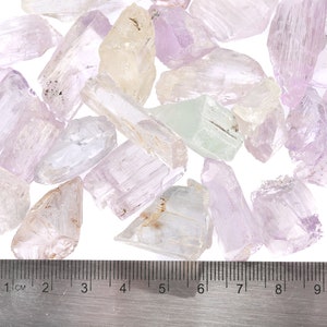 May include: A collection of pale pink, white, and green crystal points, some with a slight yellow tint, are scattered on a white surface. A ruler is placed below the crystals, showing measurements in centimeters.