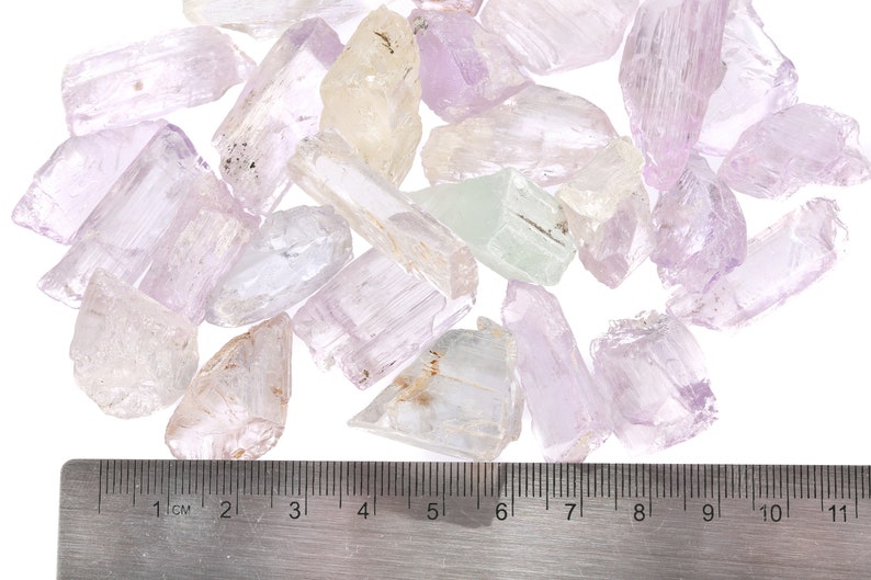 May include: A collection of pale pink, white, and green crystal points, some with a slight yellow tint, are scattered on a white surface. A ruler is placed below the crystals, showing measurements in centimeters.