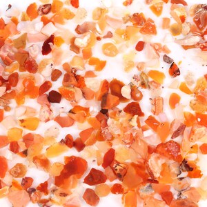 May include: A collection of small, irregularly shaped, orange and red gemstones. The gemstones are scattered on a white surface.