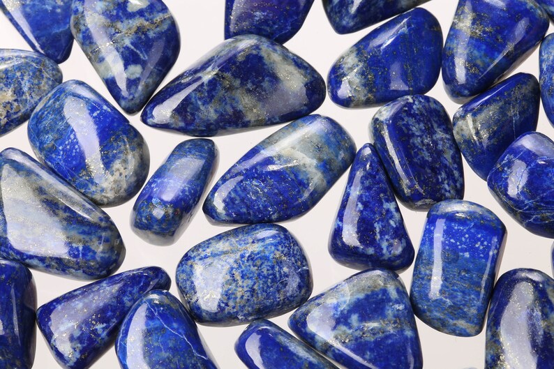 May include: A collection of tumbled lapis lazuli stones. The stones are a deep blue color with white and gold flecks. They are smooth and polished.