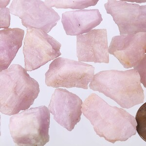 May include: A collection of pink and white rough gemstones, some with a slightly translucent appearance. The gemstones are scattered on a white surface, with a US penny for scale.