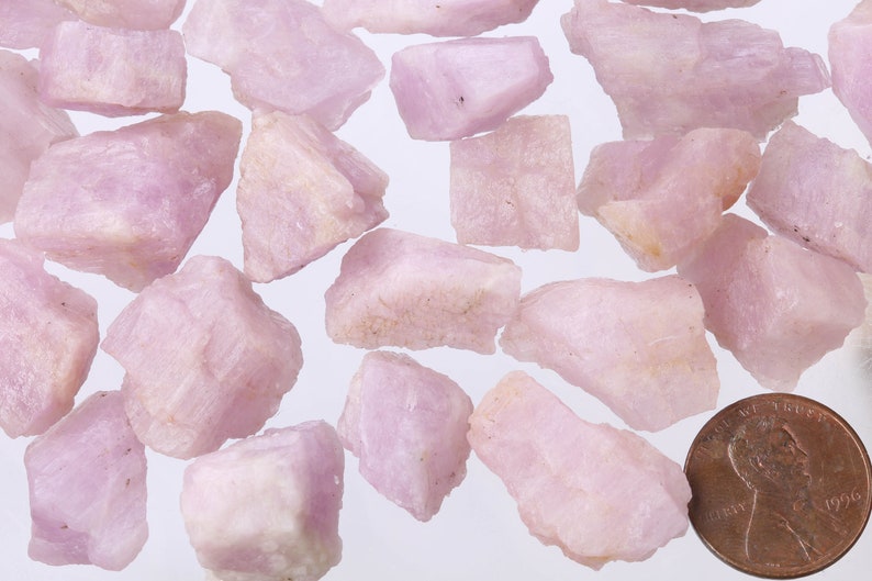 May include: A collection of pink and white rough gemstones, some with a slightly translucent appearance. The gemstones are scattered on a white surface, with a US penny for scale.