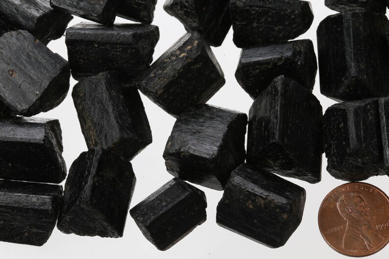 May include: A collection of black tourmaline crystals, some with a slight sheen, arranged on a white surface. A US penny is included for scale.