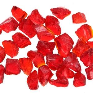 May include: A collection of red glass shards, some with a slight orange hue, scattered on a white background. The shards are irregular in shape and size, with sharp edges.