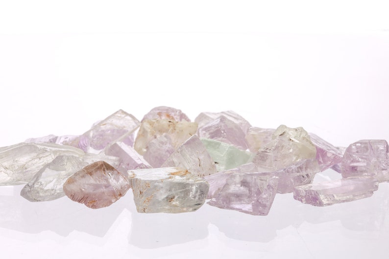 May include: A collection of pale pink, white, and green crystals with a variety of shapes and sizes. The crystals are arranged in a pile on a white surface.