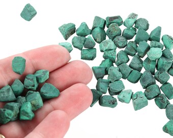 Op de afbeelding: Een hoop kleine, groene malachietstenen. Sommige stenen bevinden zich in een hand, terwijl andere verspreid liggen op een wit oppervlak.