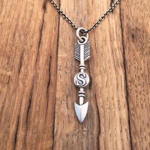 May include: Silver arrow necklace with a letter 'S' charm. The arrow is detailed with a feathered design and a pointed tip. The necklace is on a wooden background.