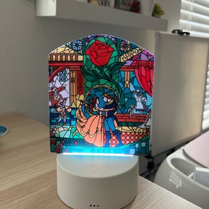 Puede incluir: Una base blanca iluminada con un diseño de vitral que representa a Bella y la Bestia de La Bella y la Bestia. El diseño de vitral incluye una rosa roja, un castillo y otros detalles. La base tiene un botón de encendido y una luz azul que brilla a través del vitral.