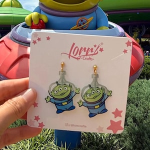 Puede incluir: Pendientes acrílicos con los Little Green Men de Toy Story. Los pendientes son transparentes con detalles en verde, azul y dorado. Se exhiben en una tarjeta blanca con el texto "Lorr's Crafts" y estrellas rosas.