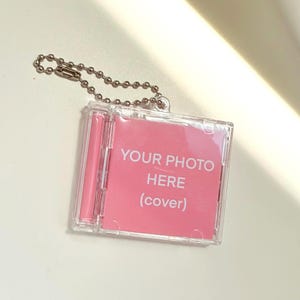 May include: A clear plastic keychain with a pink insert that reads "YOUR PHOTO HERE (cover)". The keychain has a silver ball chain attached for easy carrying. The design is simple and modern, perfect for displaying a small photo.