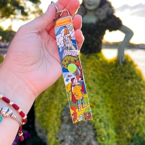 May include: A colorful keychain featuring a castle, a prince and princess, and a forest scene. The keychain is held in a hand with a red bracelet.