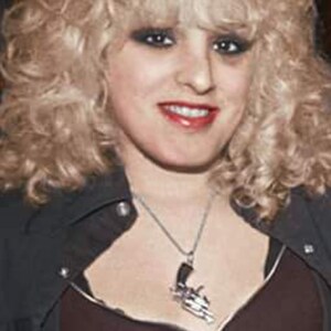 May include: A woman with blonde curly hair wearing a black leather jacket with a zipper and a silver necklace with a gun pendant.