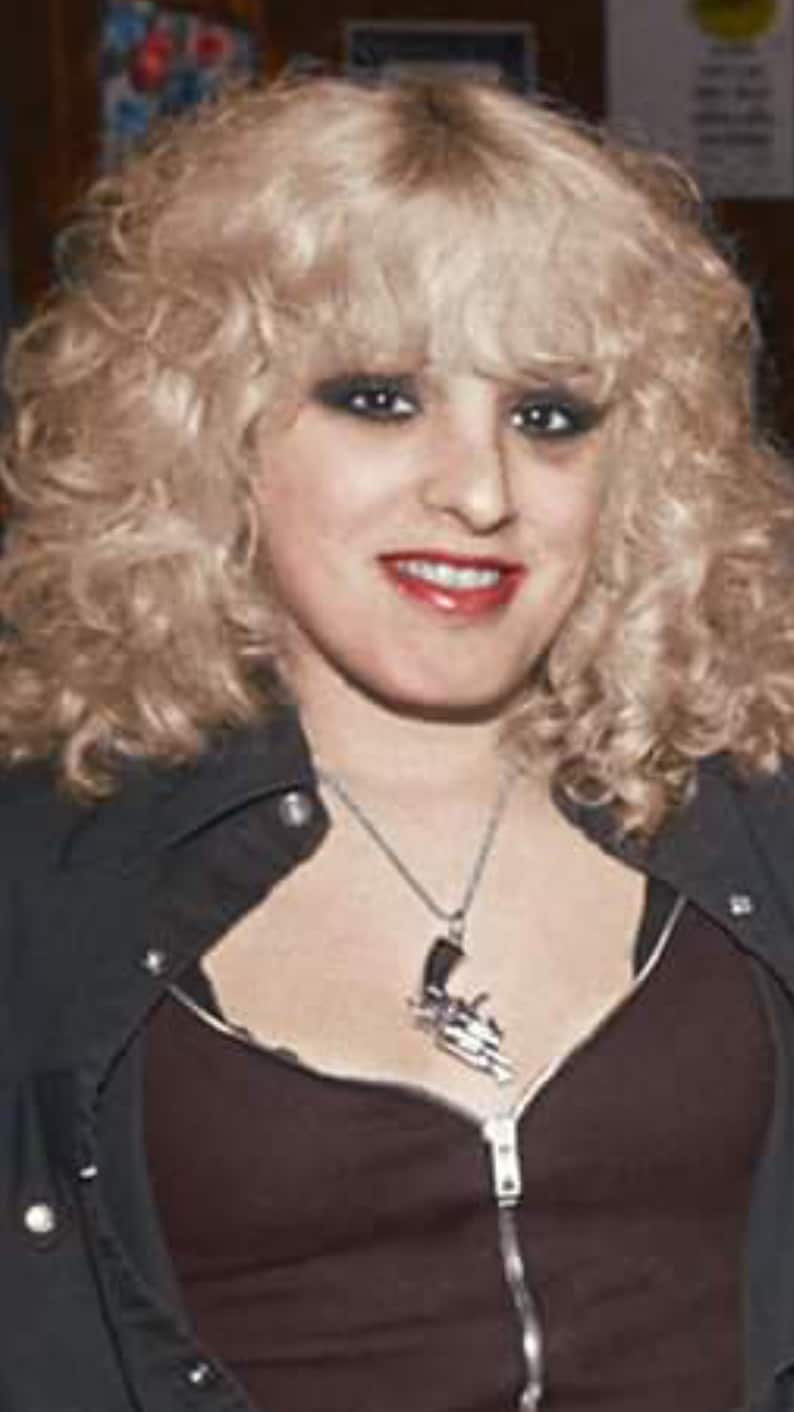 May include: A woman with blonde curly hair wearing a black leather jacket with a zipper and a silver necklace with a gun pendant.