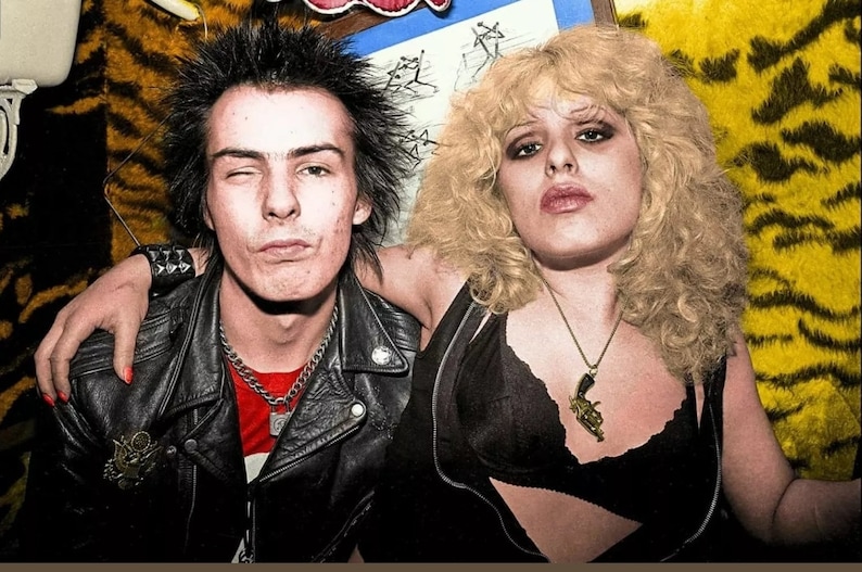Pode incluir: Uma fotografia a preto e branco de um homem e uma mulher com penteados punk rock. O homem est&aacute; a usar uma jaqueta de couro preta com uma camisa vermelha por baixo. A mulher est&aacute; a usar um top preto com um colar de ouro com um pingente de pistola. Ambos est&atilde;o a olhar para a c&acirc;mara.