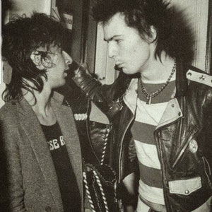 May include: Black and white photograph of two people in a dimly lit room. One person wears a striped shirt under a leather jacket, the other a blazer and a t-shirt with text. The image captures a moment in time.