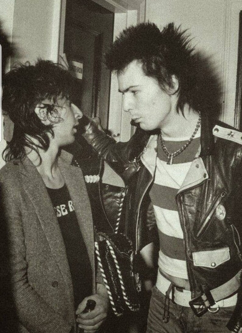 May include: Black and white photograph of two people in a dimly lit room. One person wears a striped shirt under a leather jacket, the other a blazer and a t-shirt with text. The image captures a moment in time.