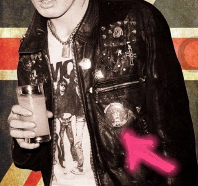 May include: Black leather jacket with metal studs and patches. The jacket is worn over a white graphic t-shirt. A glass of a light-colored beverage is held in the hand. The background has a vintage aesthetic.