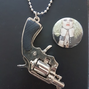 Puede incluir: Un colgante de pistola de juguete de plata en una cadena de plata. El colgante tiene un mango de plástico negro. El collar está sobre un fondo negro. Un botón redondo con la cara de una mujer está en el fondo.