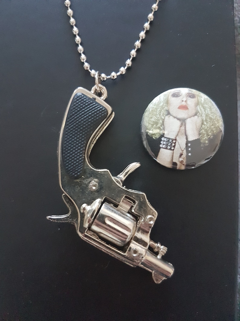 May include: A silver toy gun pendant on a silver chain necklace. The pendant has a black plastic handle. The necklace is on a black background. A round button with a woman's face on it is in the background.