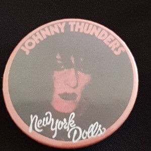 May include: A round button with a pink border and a gray center featuring a portrait of Johnny Thunders. The text "JOHNNY THUNDERS" arches across the top, and "New York Dolls" is written in cursive at the bottom.