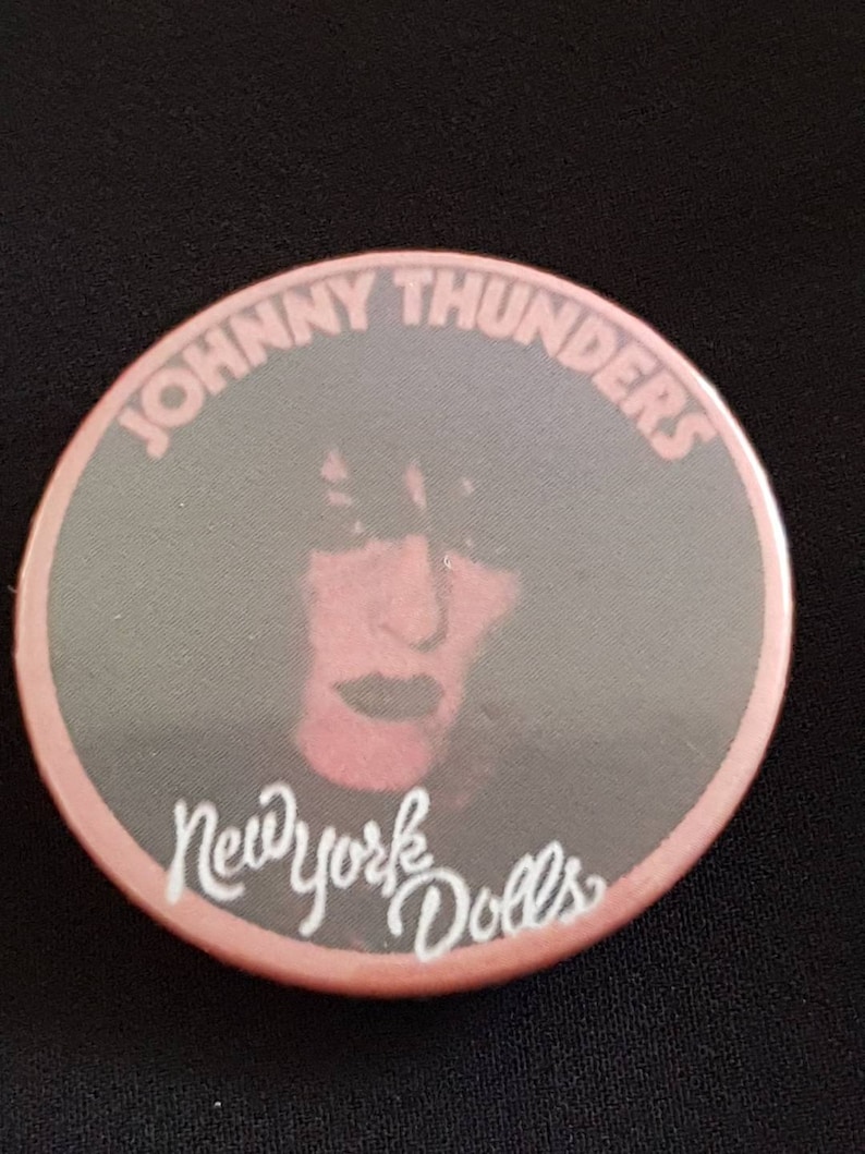 May include: A round button with a pink border and a gray center featuring a portrait of Johnny Thunders. The text "JOHNNY THUNDERS" arches across the top, and "New York Dolls" is written in cursive at the bottom.