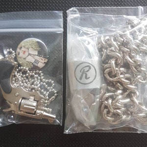 May include: Two silver necklaces in clear plastic bags. One necklace features a silver toy gun charm and a round pin with a woman's face. The other necklace features a silver chain and a silver charm with the letter "R" in a circle.