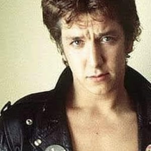 May include: A portrait of a person with dark, spiky hair and a black leather jacket. The jacket has a collar and two round pins. The person is looking directly at the camera.
