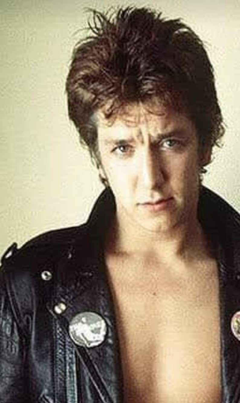May include: A portrait of a person with dark, spiky hair and a black leather jacket. The jacket has a collar and two round pins. The person is looking directly at the camera.
