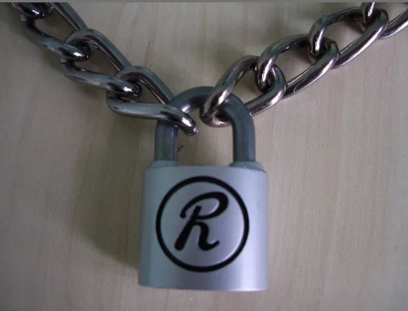 May include: A silver padlock with a silver chain attached. The padlock has a black circle with a silver letter 'R' in the center. 19" chain, & two keys.