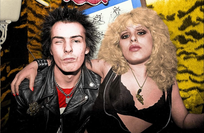 May include: A black and white photo of a couple. The man has short, spiky black hair and is wearing a black leather jacket, a red shirt, and a silver chain necklace. The woman has long, blonde hair and is wearing a black bra and a gold necklace with a gun pendant.