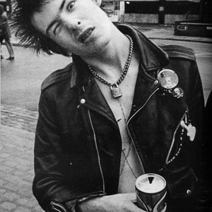 May include: Black and white photograph of a person in a leather jacket, chain necklace with a padlock pendant, and multiple pins. The person is holding a can of soda. The image captures a punk rock aesthetic.