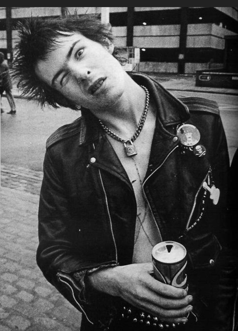 May include: Black and white photograph of a person in a leather jacket, chain necklace with a padlock pendant, and multiple pins. The person is holding a can of soda. The image captures a punk rock aesthetic.