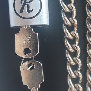 Pode incluir: Um cadeado prateado com uma letra "R" preta na frente, uma chave prateada e uma corrente prateada.