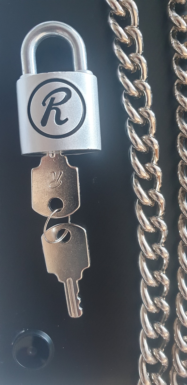 Pode incluir: Um cadeado prateado com uma letra "R" preta na frente, uma chave prateada e uma corrente prateada.