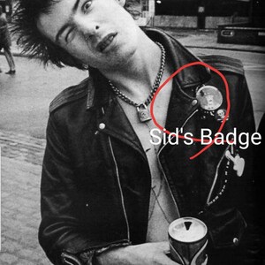 May include: Black and white image of a person wearing a black leather jacket with a chain necklace and a padlock pendant. The jacket has a round badge and other pins. The person is holding a can.