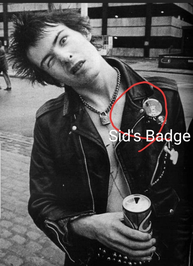 May include: Black and white image of a person wearing a black leather jacket with a chain necklace and a padlock pendant. The jacket has a round badge and other pins. The person is holding a can.
