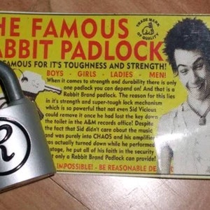 Pode incluir: Um cadeado prateado com uma letra "R" preta na frente. O cadeado &eacute; anunciado como "O famoso cadeado Rabbit" e &eacute; descrito como "resistente" e "forte". O an&uacute;ncio apresenta uma foto em preto e branco de um homem com um penteado punk rock e uma camiseta branca. O texto do an&uacute;ncio diz "Usado com orgulho por Sid Vicious do grupo pop Sex Pistols!"
