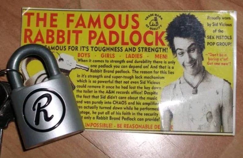 Pode incluir: Um cadeado prateado com uma letra "R" preta na frente. O cadeado &eacute; anunciado como "O famoso cadeado Rabbit" e &eacute; descrito como "resistente" e "forte". O an&uacute;ncio apresenta uma foto em preto e branco de um homem com um penteado punk rock e uma camiseta branca. O texto do an&uacute;ncio diz "Usado com orgulho por Sid Vicious do grupo pop Sex Pistols!"