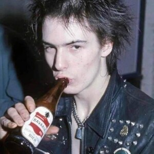 May include: A person drinking from a brown Bavaria Dortmunder Bier bottle. The person is wearing a black leather jacket with silver studs and a silver chain necklace. A round pin with text is visible on the jacket.