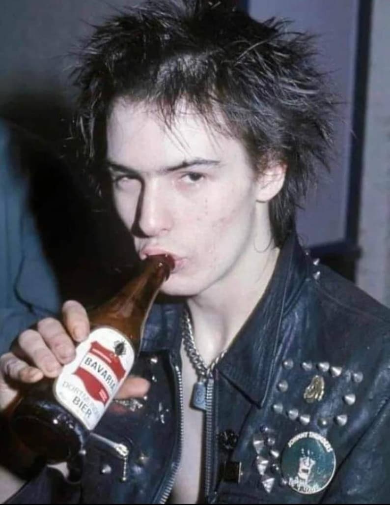 May include: A person drinking from a brown Bavaria Dortmunder Bier bottle. The person is wearing a black leather jacket with silver studs and a silver chain necklace. A round pin with text is visible on the jacket.